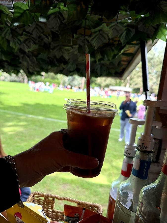 An iced coffee in a clear cup held up in front of a sunny village green
